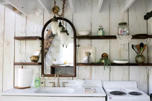 A cozy kitchen with white wooden walls, shelves displaying dishes, a mirror, and a vintage stove.