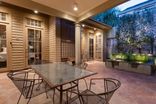 A cozy outdoor patio with a glass table and metal chairs, surrounded by greenery and warm lighting.