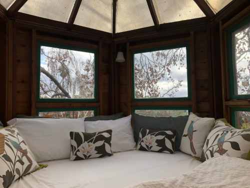 Cozy nook with a bed surrounded by large windows, offering views of trees and a cloudy sky. Decorative pillows add comfort.