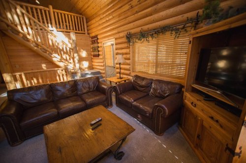 Cozy log cabin living room with leather sofas, a wooden coffee table, and warm lighting from large windows.