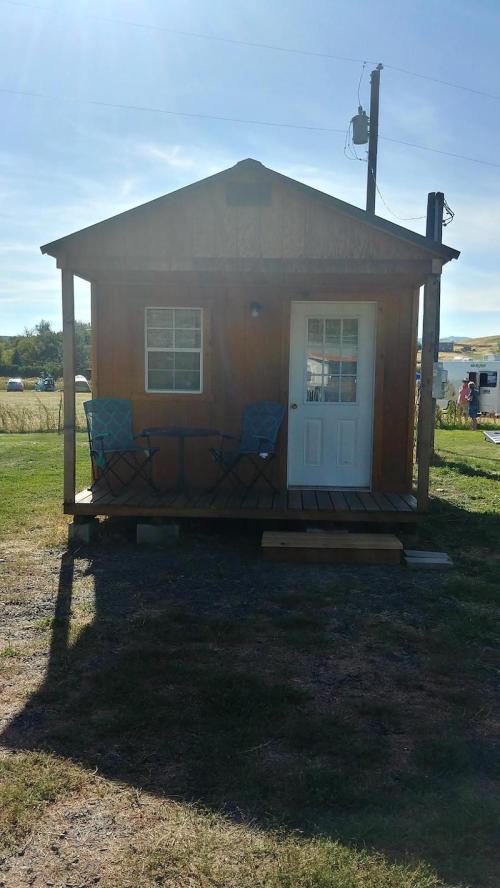 A small wooden cabin with a porch, two blue chairs, and a door, set in a grassy area under a clear sky.