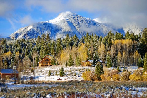 A snowy mountain landscape with evergreen trees, colorful autumn foliage, and cozy cabins under a blue sky.
