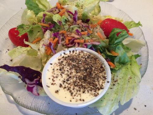 A fresh salad with mixed greens, carrots, and tomatoes, served with a small bowl of creamy dressing topped with pepper.