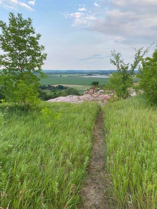 A grassy path leads to a rocky outcrop, with lush greenery and a scenic view of fields and distant hills.