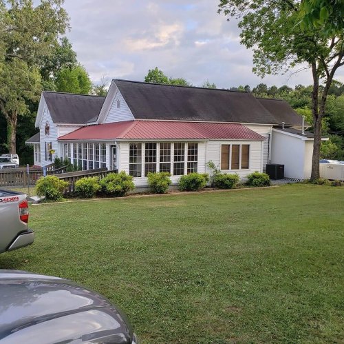 A white house with a red roof, surrounded by greenery and a lawn, under a partly cloudy sky.