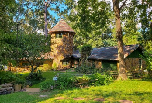 A whimsical stone house with a tower, surrounded by lush greenery and trees in a sunny garden setting.