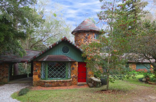 A charming stone cottage with a red door and turret, surrounded by greenery and a gravel path.