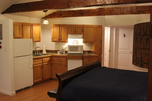 Cozy kitchen and bedroom area with wooden beams, featuring a fridge, microwave, and a bed with a dark blue cover.