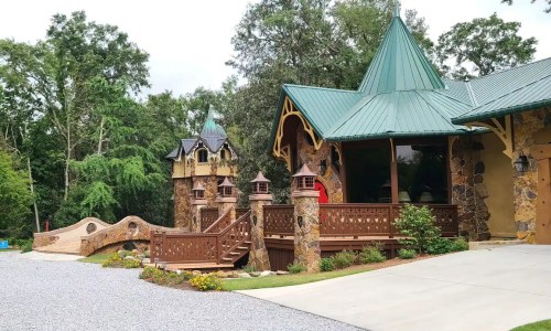 A whimsical stone house with a green metal roof, surrounded by trees and featuring a decorative bridge and garden.