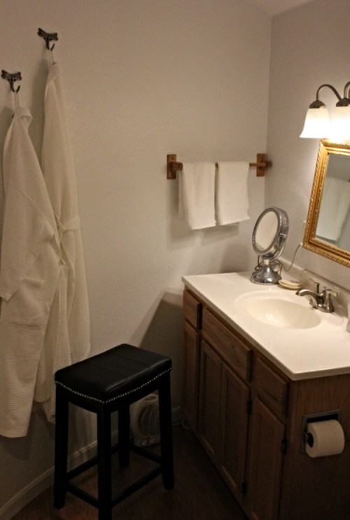 A clean bathroom with a sink, mirror, towels, a stool, and bathrobes hanging on hooks.