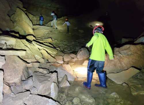 A group of people exploring a dark cave, navigating rocky terrain with headlamps and wearing waterproof boots.