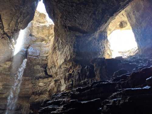 A cavern with two openings, sunlight streaming in, and a waterfall cascading down rocky walls.