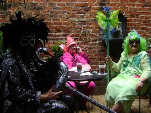 Three people in colorful costumes sit at a table in a brick courtyard, surrounded by greenery and drinks.