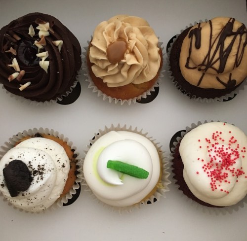 A box of six assorted cupcakes with various toppings, including chocolate, vanilla, and peanut butter flavors.