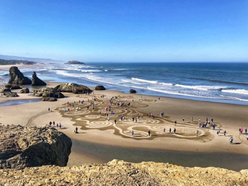 A beach scene with people creating intricate sand patterns, surrounded by rocks and ocean waves under a clear blue sky.