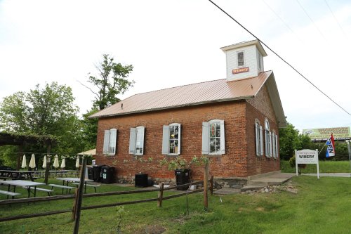 A rustic brick winery building with a metal roof, surrounded by greenery and outdoor seating.
