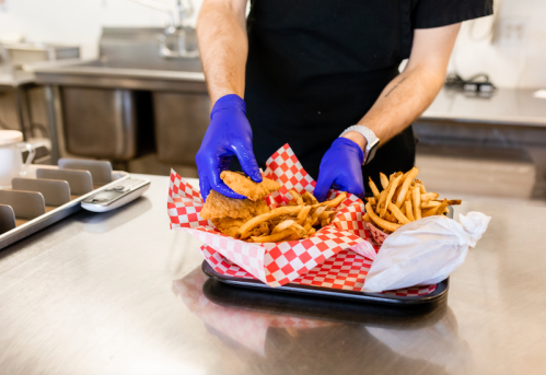 A person in gloves serves fried chicken and fries in a red-checkered basket on a stainless steel countertop.