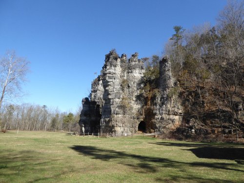 Rock formations rise against a clear blue sky, surrounded by sparse trees and a grassy area in the foreground.