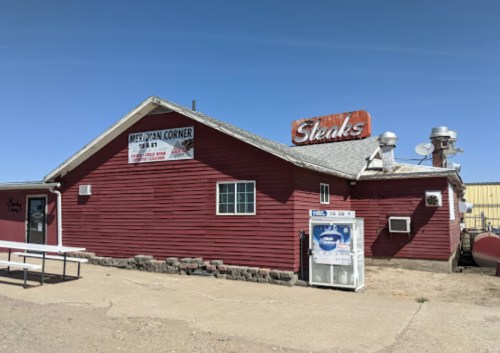 Red wooden building with a sign reading "Steaks" and "Meridian Corner," set against a clear blue sky.