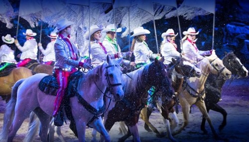 A group of riders in white hats and sequined outfits perform on horseback under colorful lights and umbrellas.