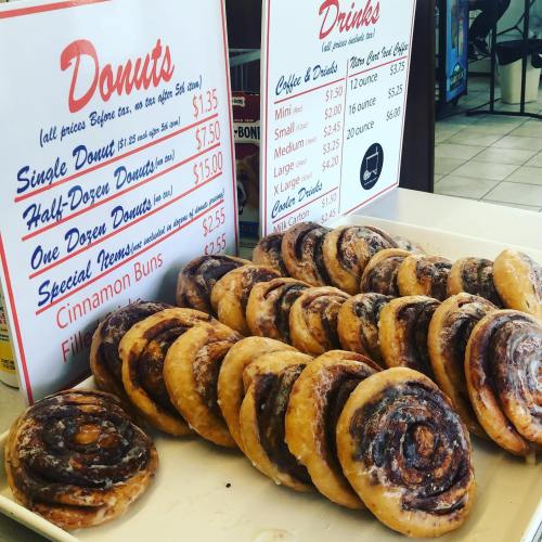 A display of donuts and cinnamon buns with a price list for donuts and drinks in a shop setting.