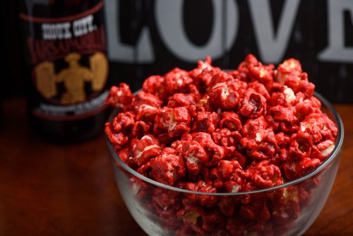 A bowl of bright red popcorn sits on a wooden surface, with a bottle of soda in the background.