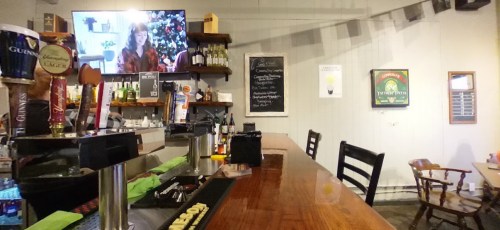 A cozy bar with a wooden counter, shelves of drinks, and a TV showing a woman by a Christmas tree.