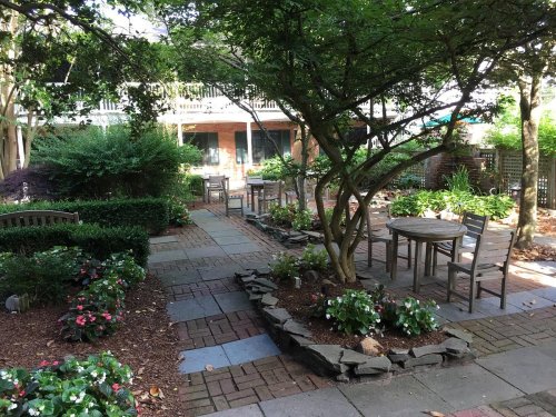 A serene garden with stone pathways, tables, and chairs surrounded by lush greenery and blooming flowers.