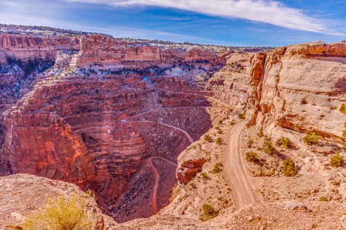 A winding dirt road curves through a deep canyon with layered red rock formations under a blue sky.