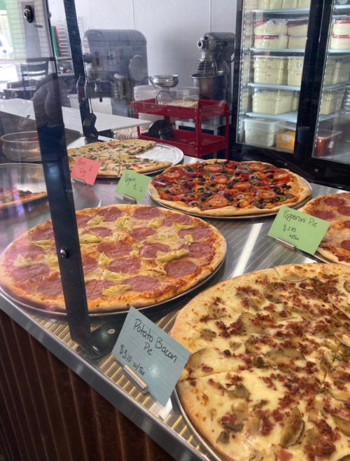A display case with various pizzas, including pepperoni, veggie, and potato bacon, each labeled with prices.