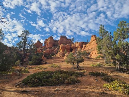 Rocky formations rise against a blue sky with scattered clouds, surrounded by trees and shrubs in a natural landscape.
