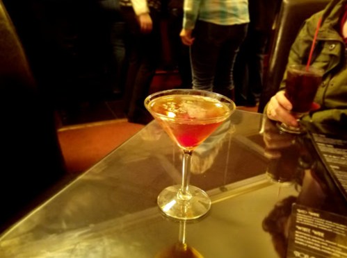 A cocktail in a martini glass sits on a table, with blurred figures in the background.