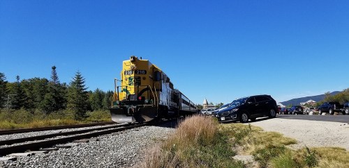 A yellow train approaches a parking area beside the tracks, with trees and mountains in the background under a clear blue sky.