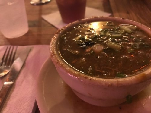 A bowl of soup with green onions, sitting on a table with utensils and a drink in the background.
