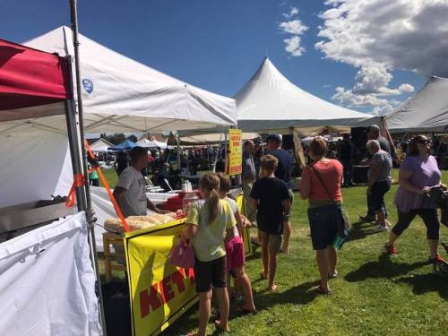 A busy outdoor market scene with people browsing tents and a vendor selling food under a sunny sky.