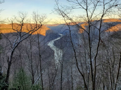 A scenic view of a river winding through a valley, framed by bare trees and bathed in warm sunset light.