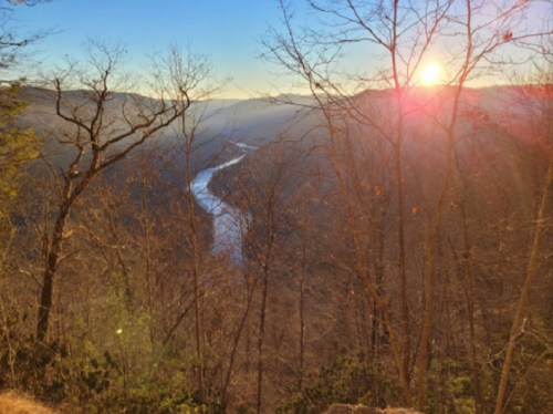 A scenic view of a river winding through a valley, framed by bare trees and a sunset in the background.