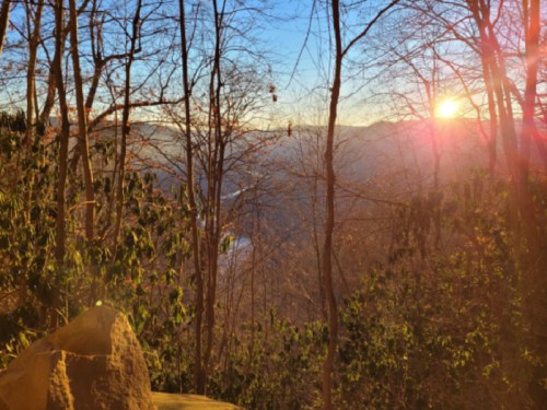 Sunset over a mountainous landscape, framed by bare trees and soft sunlight filtering through the branches.