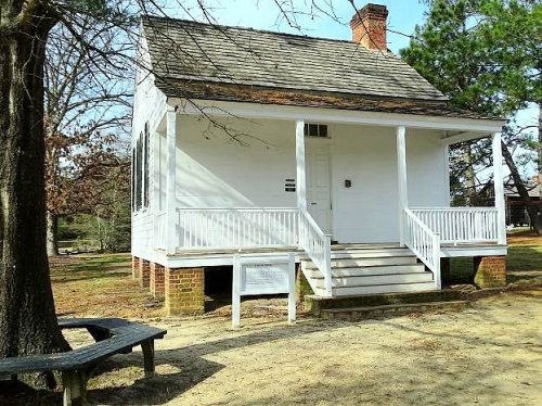 A small white house with a porch, surrounded by trees and a sandy area, featuring a brick foundation.
