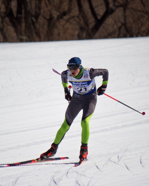 A skier in a blue cap and gray outfit races down a snowy slope, focused and determined.