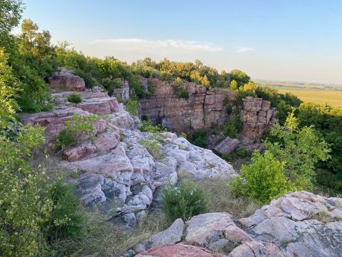 Rocky cliffside with greenery and trees, overlooking a valley under a clear blue sky.