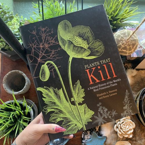 A hand holds a book titled "Plants That Kill," featuring a green illustration of a poppy on the cover, surrounded by plants.