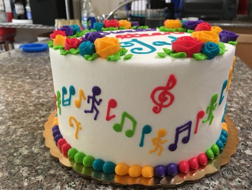 A colorful cake decorated with musical notes, dancing figures, and vibrant roses on a speckled countertop.
