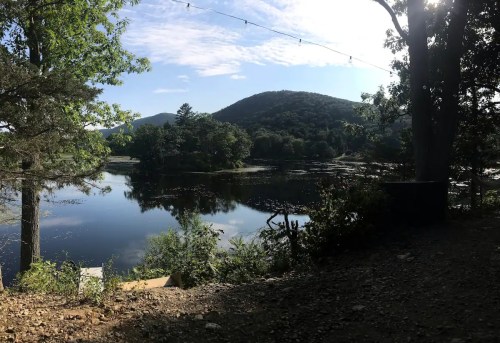 A serene lake surrounded by trees and mountains, reflecting the sky and sunlight on a calm day.