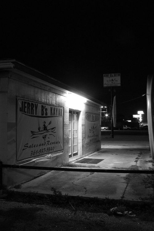 A dimly lit storefront for "Jerry B's Kayak" at night, featuring rental signs and a nearby illuminated sign.