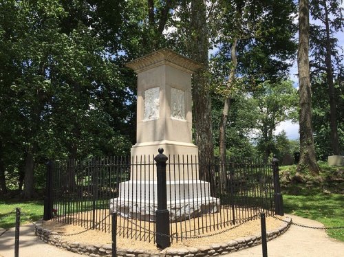 A stone monument surrounded by a black fence, set in a green park with trees and blue sky in the background.