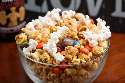 A glass bowl filled with popcorn mixed with colorful candy-coated chocolates and a dark beverage in the background.