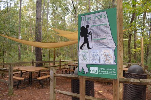 A sign about hiking trails at a nature center, with picnic tables and shaded areas in a wooded setting.