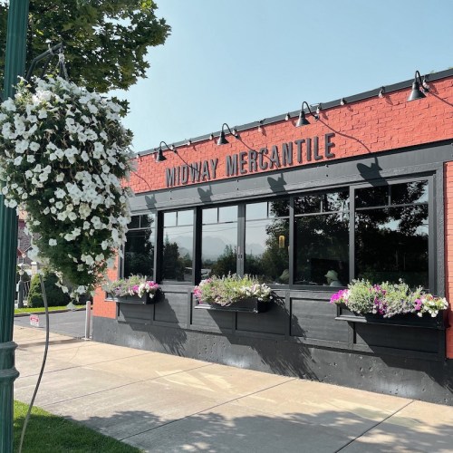 Exterior of Midway Mercantile, a red brick building with large windows and flower boxes, surrounded by greenery.