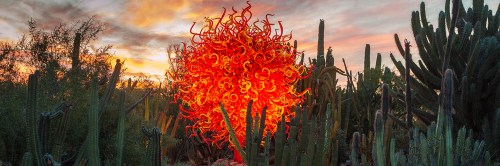 A vibrant glass sculpture resembling a fiery sun amidst cacti under a colorful sunset sky.